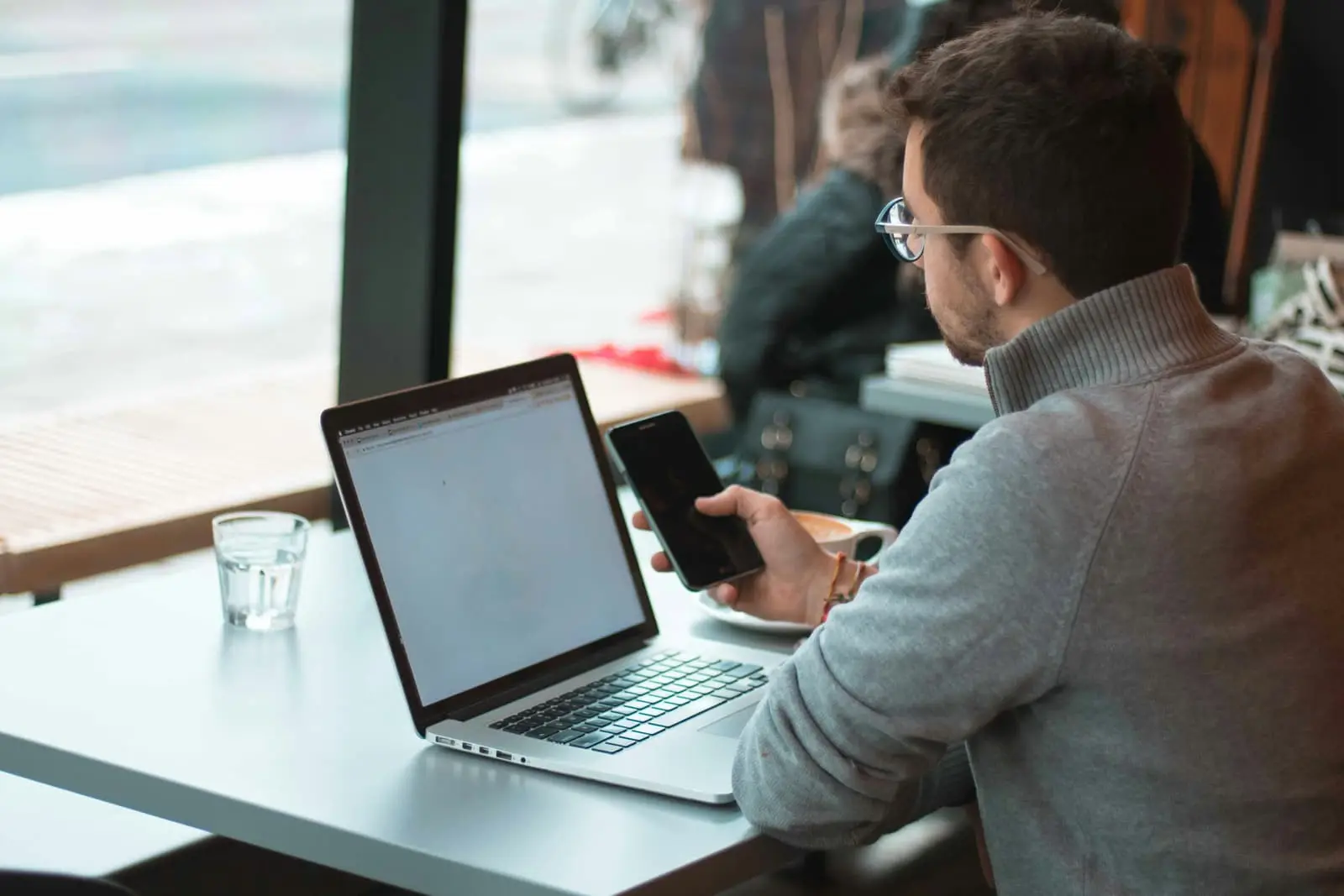 Man seated at a table with a laptop and smartphone near a window
