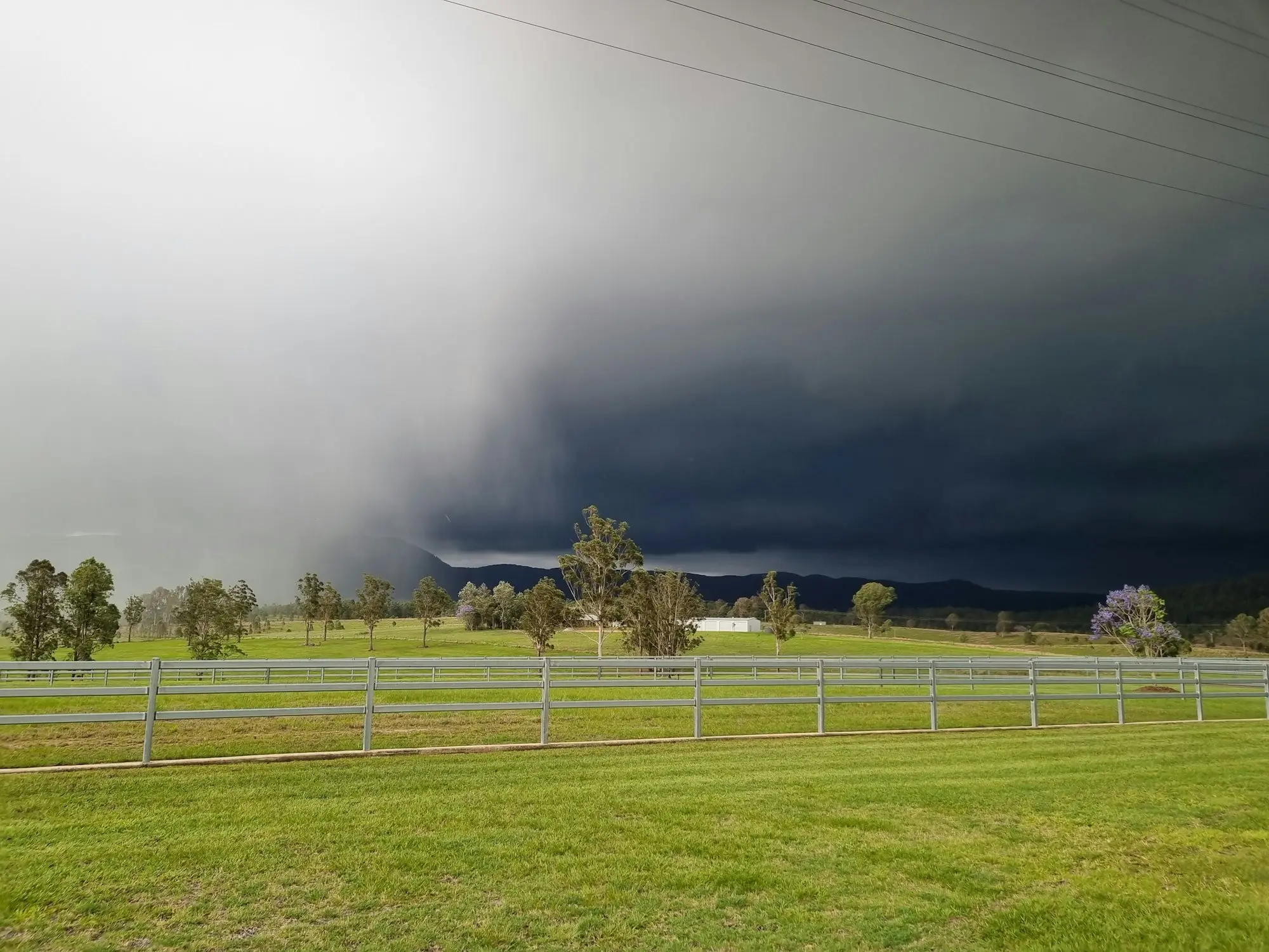 Storm clouds building over a green Australian farm paddock with a white post-and-rail fence and gum trees